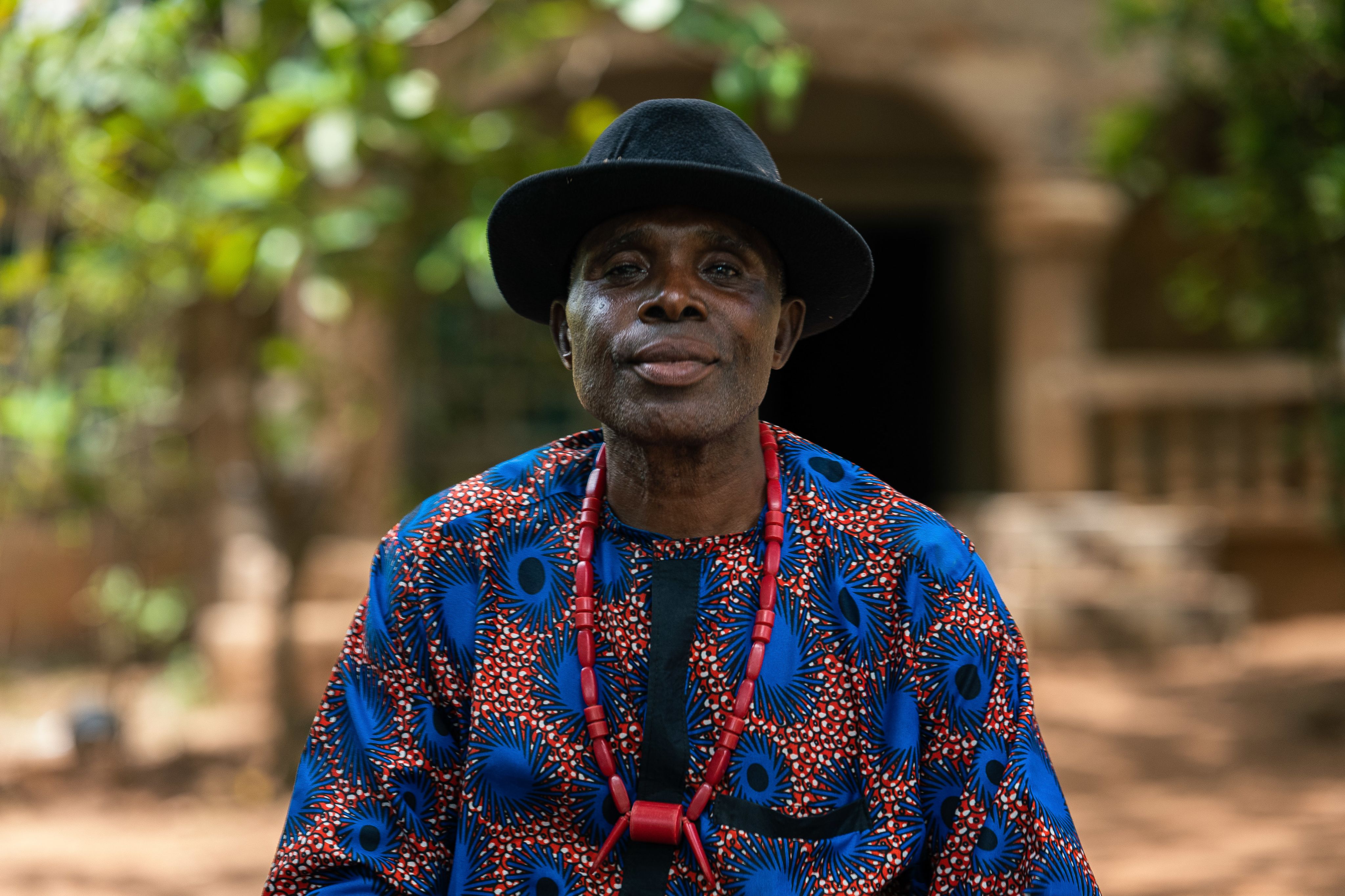 Chief Cosmos, a man in patterned clothing and a black hat, sits outside his house